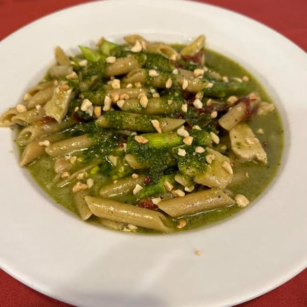 Penne pasta with green pesto sauce, chicken, asparagus, and chopped nuts on a white plate.