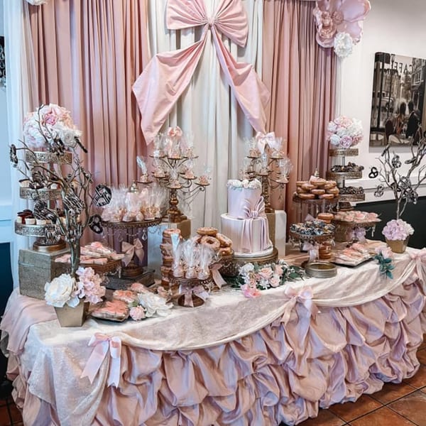 A lavish dessert table with pink and white pastries, cakes, and decorations against pink drapes.
